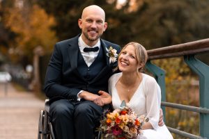 Hochzeitsfoto eines Paares in Heilbronn – Der Bräutigam im Anzug sitzt im Rollstuhl und hält die Hand seiner Braut, die ein wunderschönes Blumenbouquet hält. Beide lachen glücklich auf einer Brücke im Herbst, umgeben von bunten Bäumen. Perfekte Hochzeitsfotografie für Paare in Heilbronn und Umgebung.