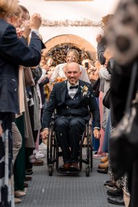 Brautpaar verlässt die Zeremonie, der Bräutigam sitzt im Rollstuhl, trägt einen eleganten Anzug mit Fliege und eine Blumenschmucknadel. Um ihn herum jubeln Gäste und Seifenblasen schweben in der Luft.