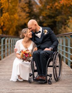 Hochzeitsfotograf Heilbronn – Authentisches Brautpaar-Shooting auf einer Holzbrücke. Braut und Bräutigam im Rollstuhl lachen gemeinsam in der Natur