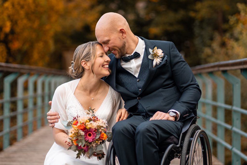 Brautpaar posiert glücklich auf einer Brücke in der Natur, die Braut hält einen Blumenstrauß in den Händen, während der Bräutigam im Rollstuhl sanft seine Stirn an ihre legt. Das herbstliche Umfeld mit Bäumen im Hintergrund verleiht der Szene Wärme und Romantik.