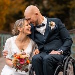 Brautpaar posiert glücklich auf einer Brücke in der Natur, die Braut hält einen Blumenstrauß in den Händen, während der Bräutigam im Rollstuhl sanft seine Stirn an ihre legt. Das herbstliche Umfeld mit Bäumen im Hintergrund verleiht der Szene Wärme und Romantik.