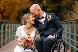 Brautpaar posiert glücklich auf einer Brücke in der Natur, die Braut hält einen Blumenstrauß in den Händen, während der Bräutigam im Rollstuhl sanft seine Stirn an ihre legt. Das herbstliche Umfeld mit Bäumen im Hintergrund verleiht der Szene Wärme und Romantik.