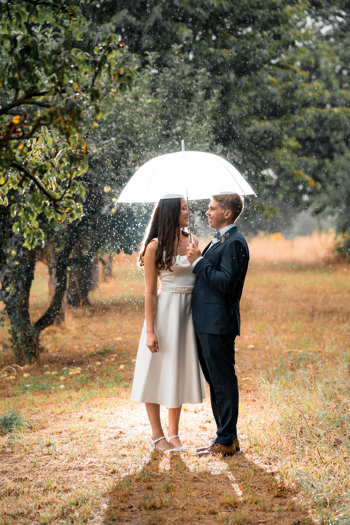 Lucia und Andi stehen einander unter einem transparenten Regenschirm im strömenden Regen gegenüber