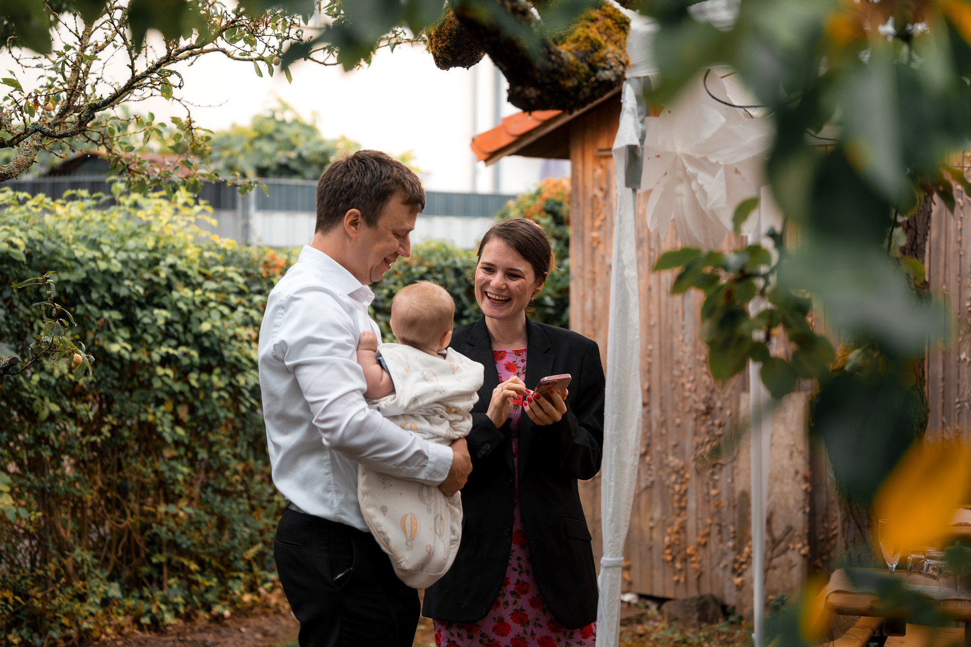 Ein Hochzeitsgast mit Baby lacht im Gespräch mit einer weiteren Person bei der Gartenfeier